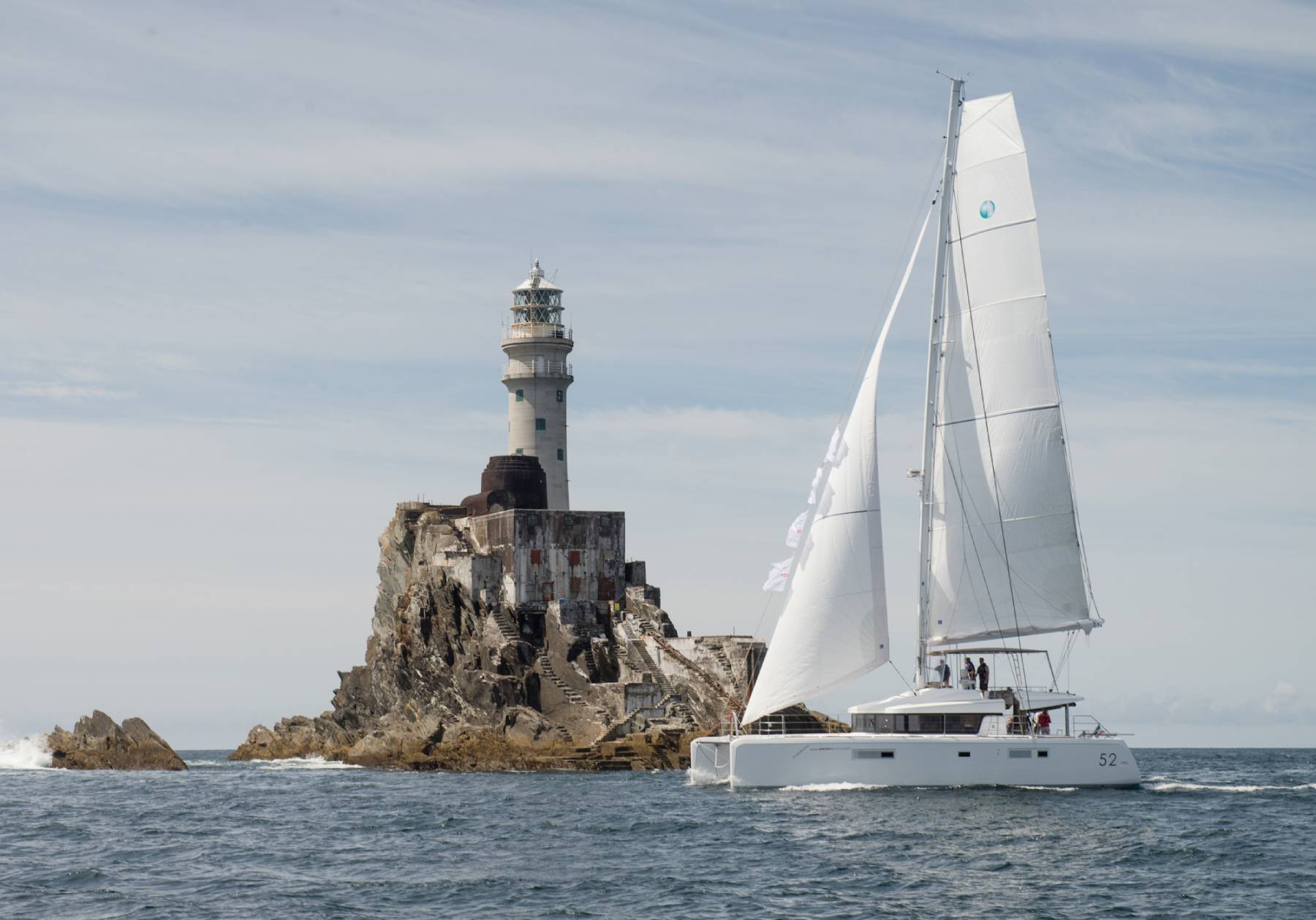 A catamaran sailboat sailing past a lighthouse located on a small rocky island. during a clear day.